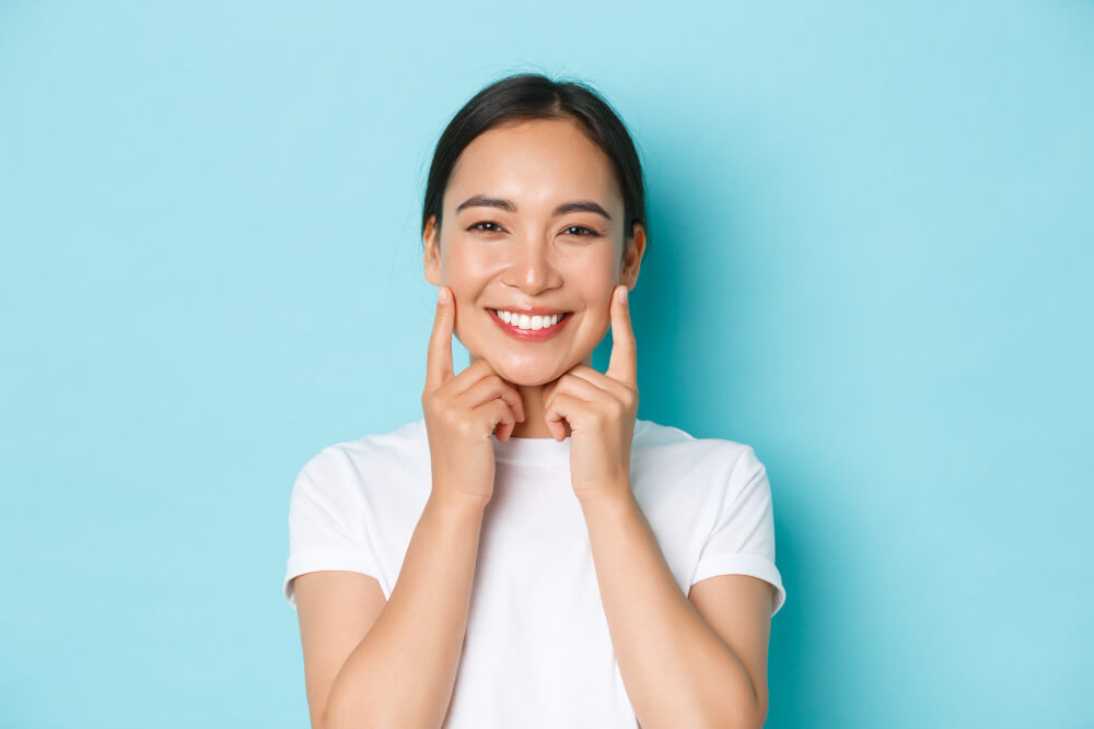 a woman in white shirt smiling brightly while pointing to her cheeks