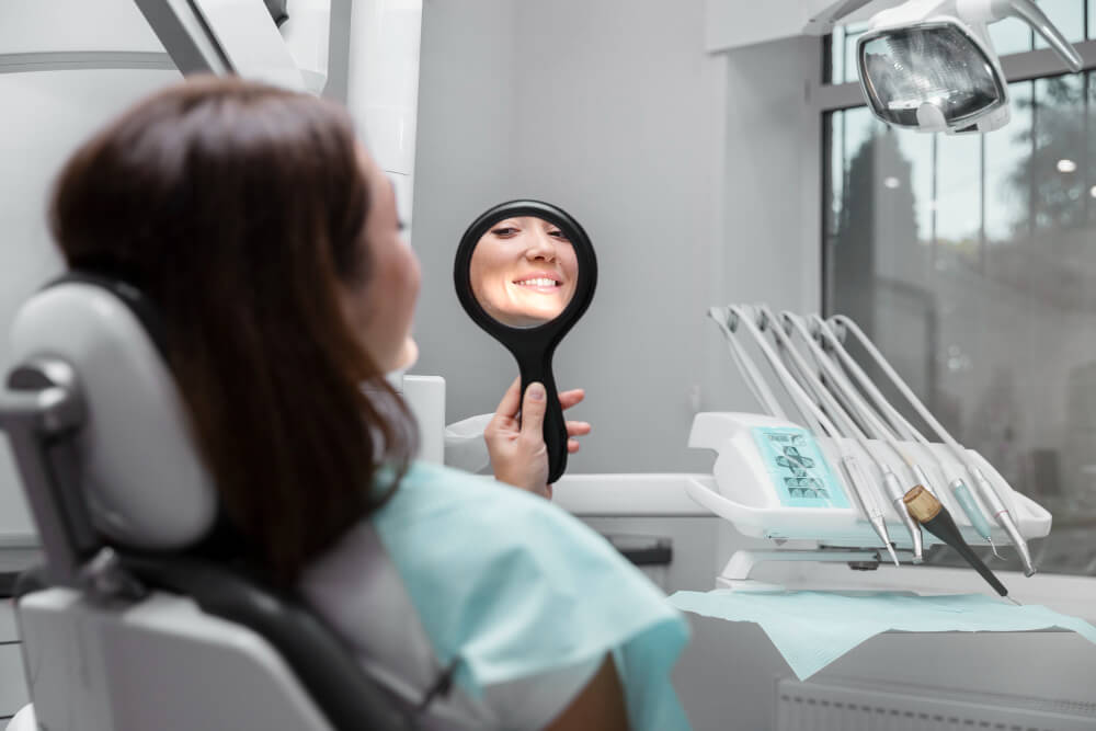 a woman at the dental chair looking at her teeth through a small mirror